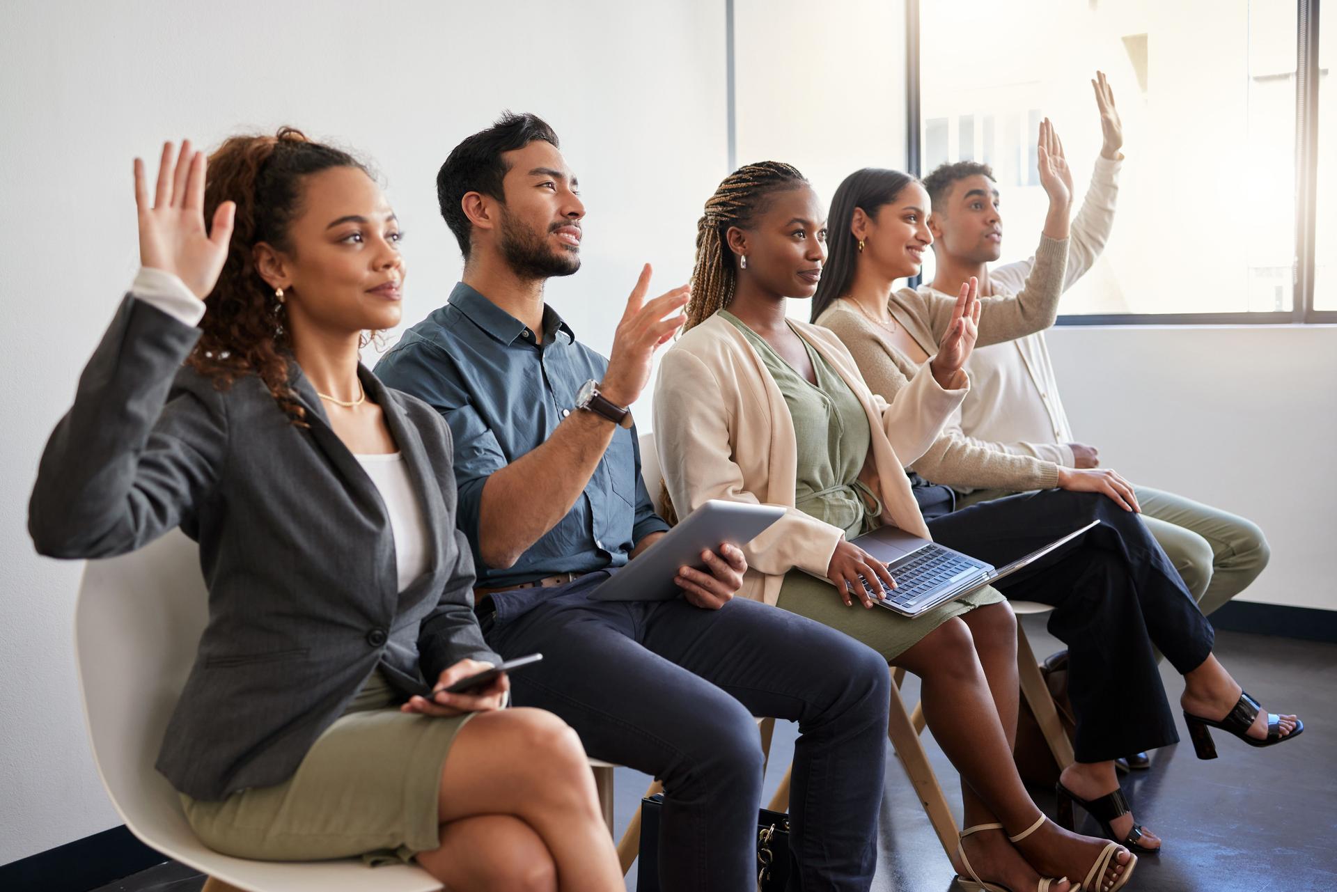 Shot of a group of young businesspeople raising their hands while sitting in a row in a modern office